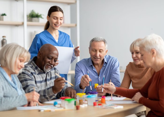 Happy multiracial elderly men and women sitting around table and drawing at nursing home, doing arts and crafts together, young lady nurse assisting group of senior people, copy space